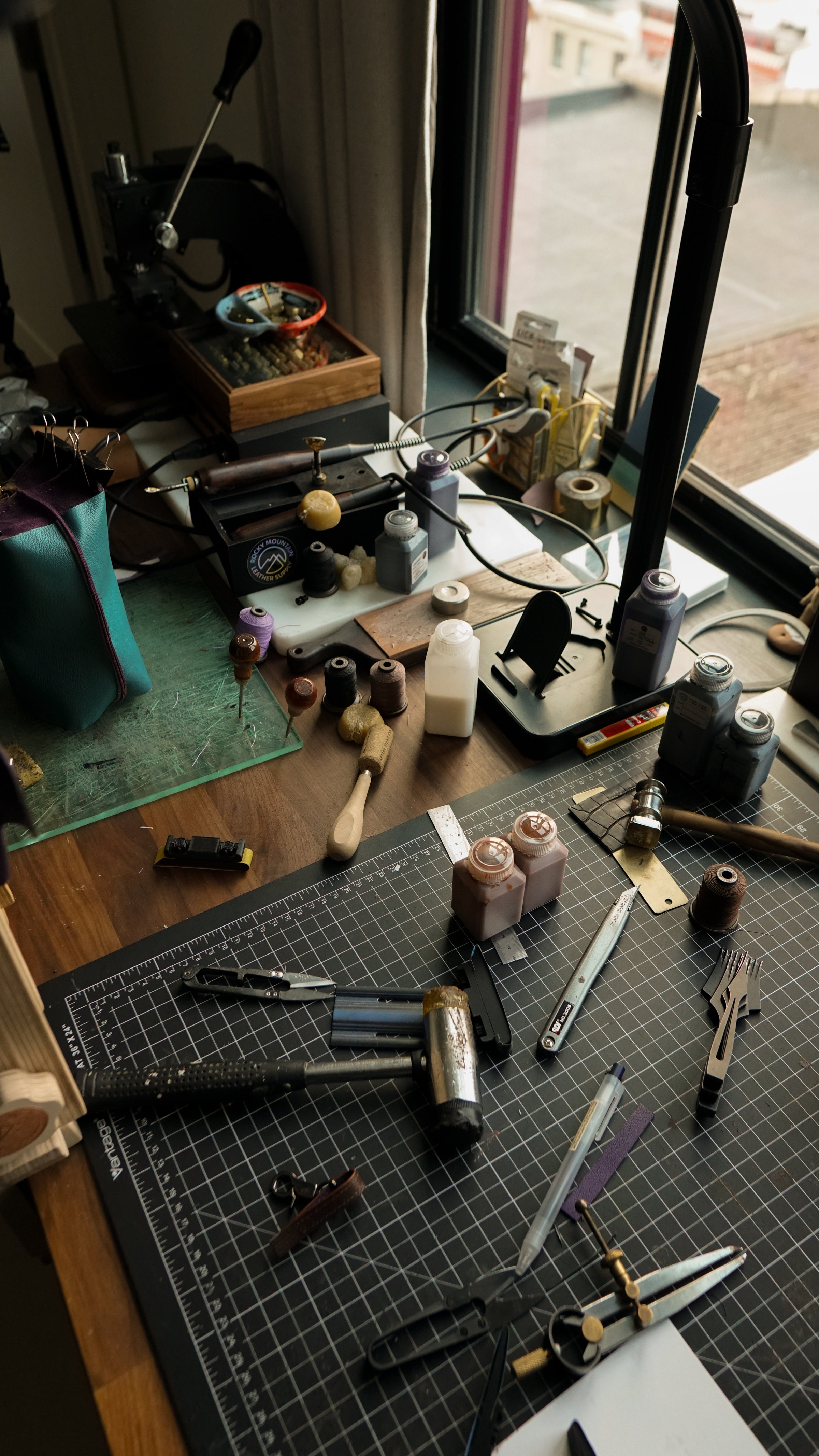 A cluttered craft workbench with various tools, small bottles, brushes, and a black gridded cutting mat in the foreground, set in a room near a window.